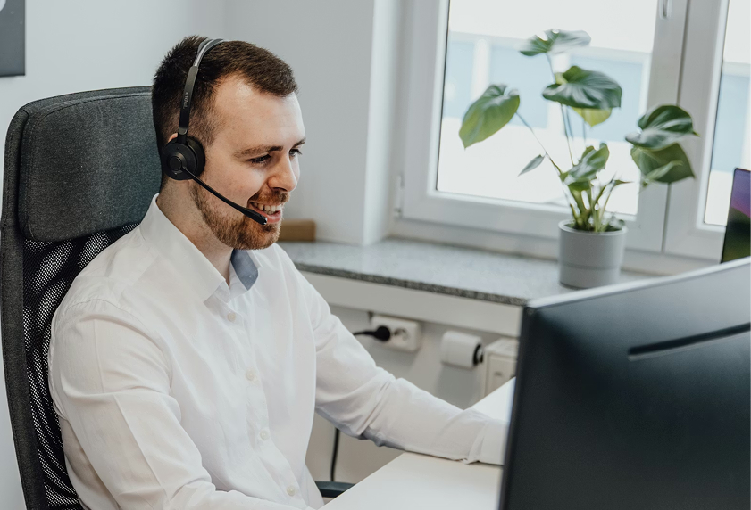 A smiling man wearing a headset and white shirt sits at a desk in a bright office, engaged in a video call or customer service interaction on his computer.